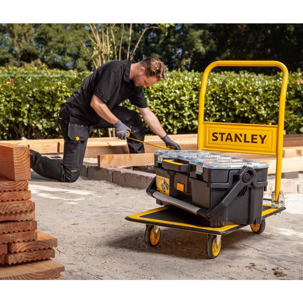 Een man meet hout naast een gele Stanley gereedschapswagen met gereedschapskist.