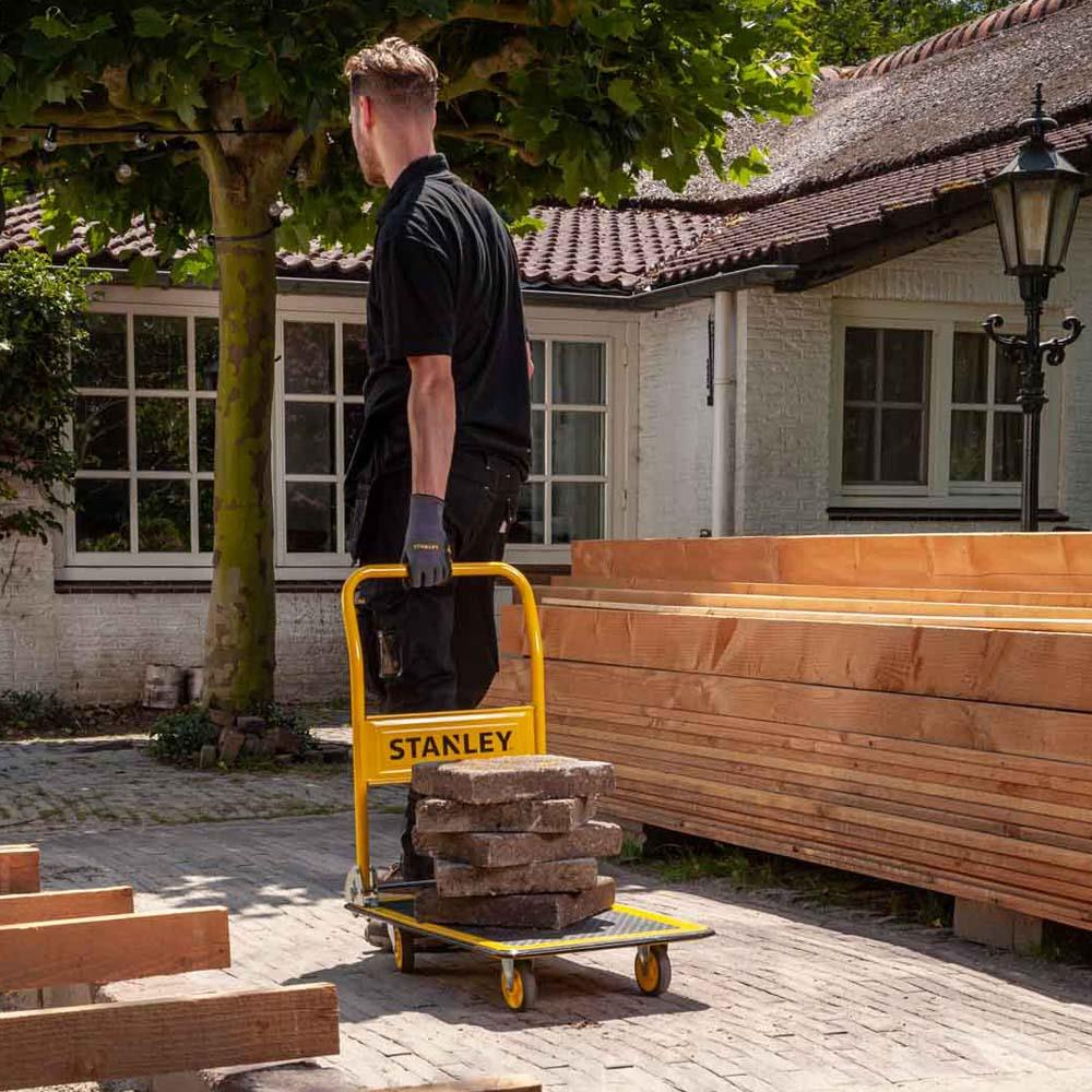 Een man duwt een gele steekwagen met stenen voor een huis met houtstapels.