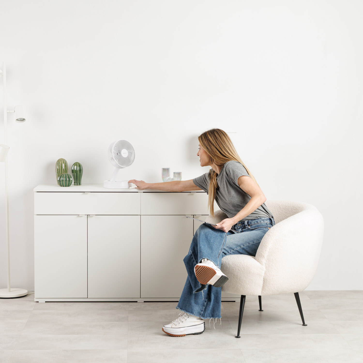 Una mujer con jeans se sienta en un sillón operando un ventilador en un mueble blanco.