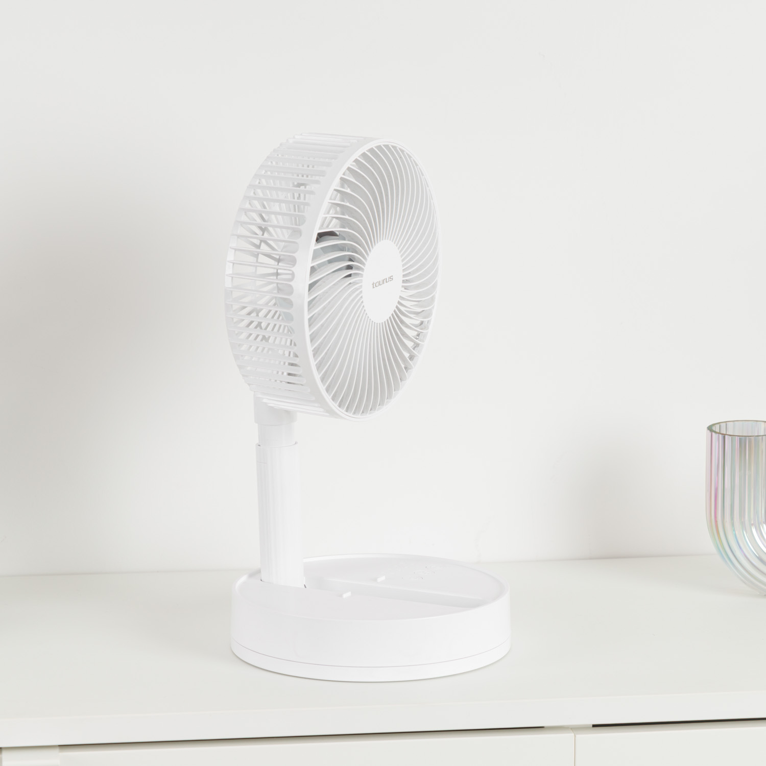 Un ventilador blanco plegable está sobre una mesa blanca junto a un jarrón de cristal de colores.