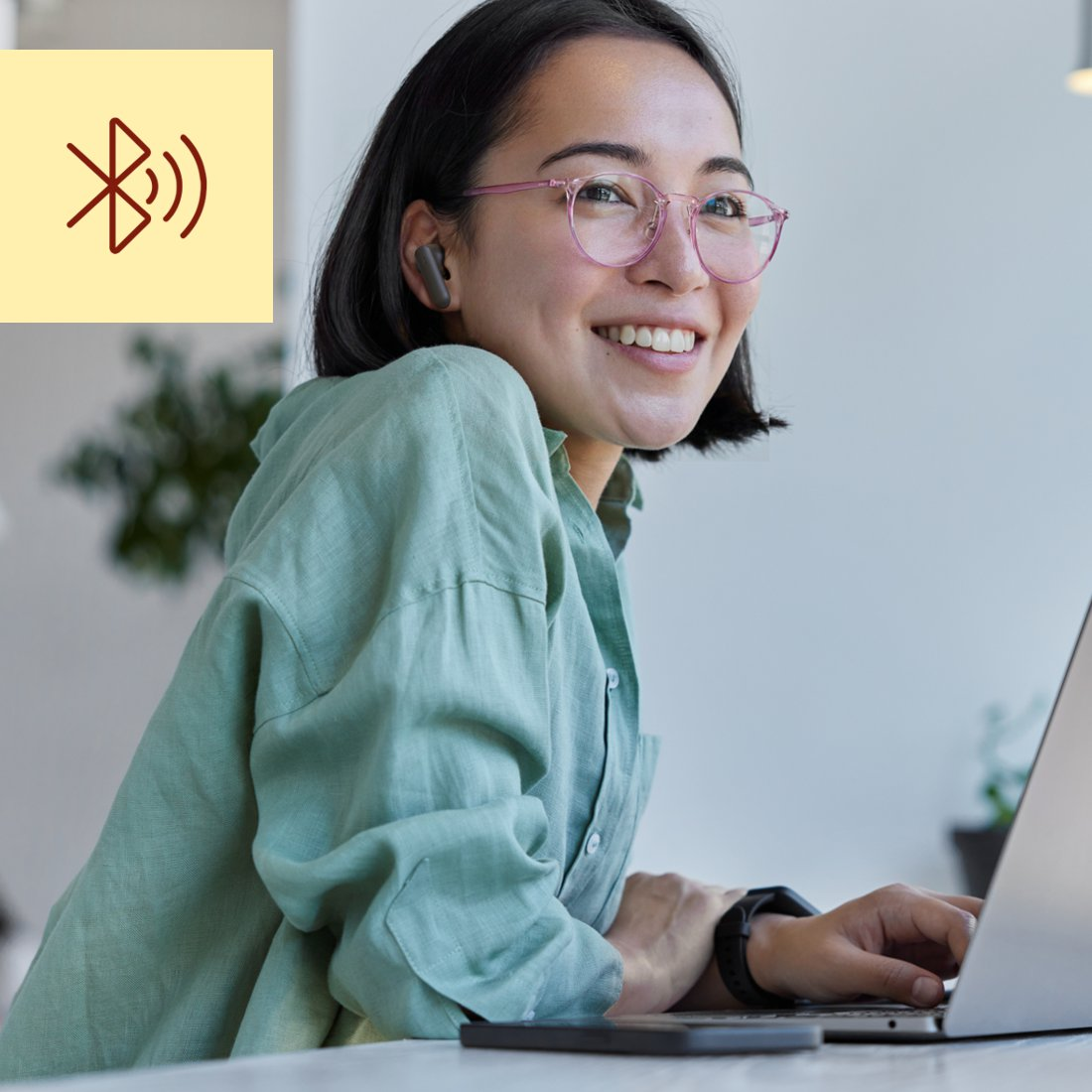 Donna con cuffie e occhiali sorride davanti al laptop con icona Bluetooth.