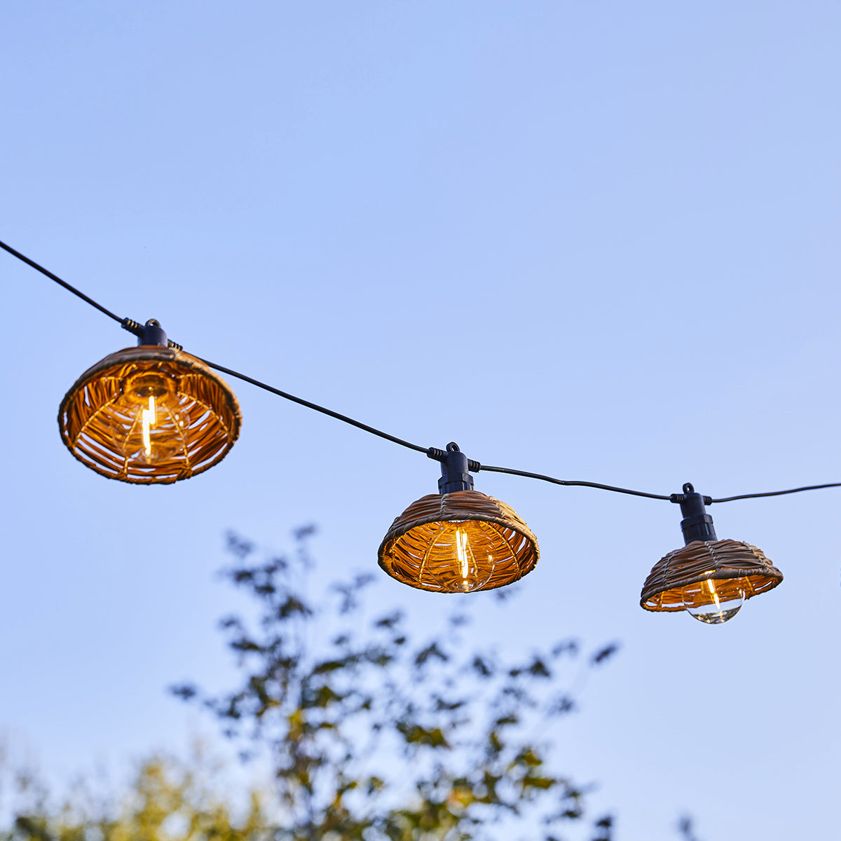 Braune Lampen mit Rattangeflecht hängen an einem Kabel vor blauem Himmel.