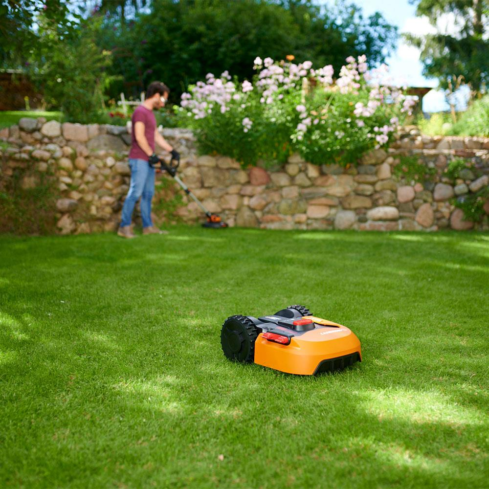 Ein orangefarbener Mähroboter steht auf grünem Rasen, im Hintergrund mäht ein Mann mit einem Trimmer.