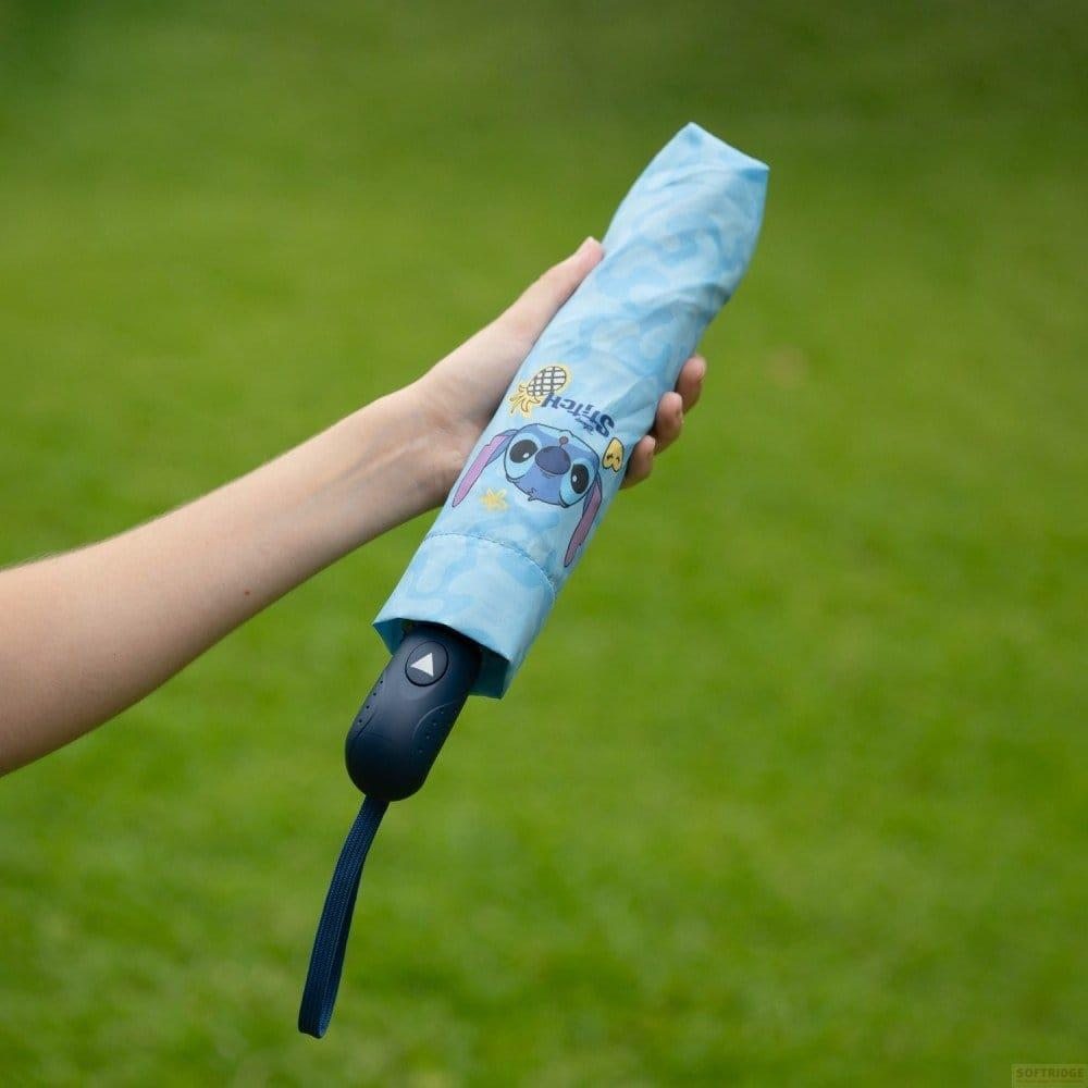 Una mano sostiene un paraguas azul cerrado con el estampado de Stitch, sobre un fondo verde borroso.