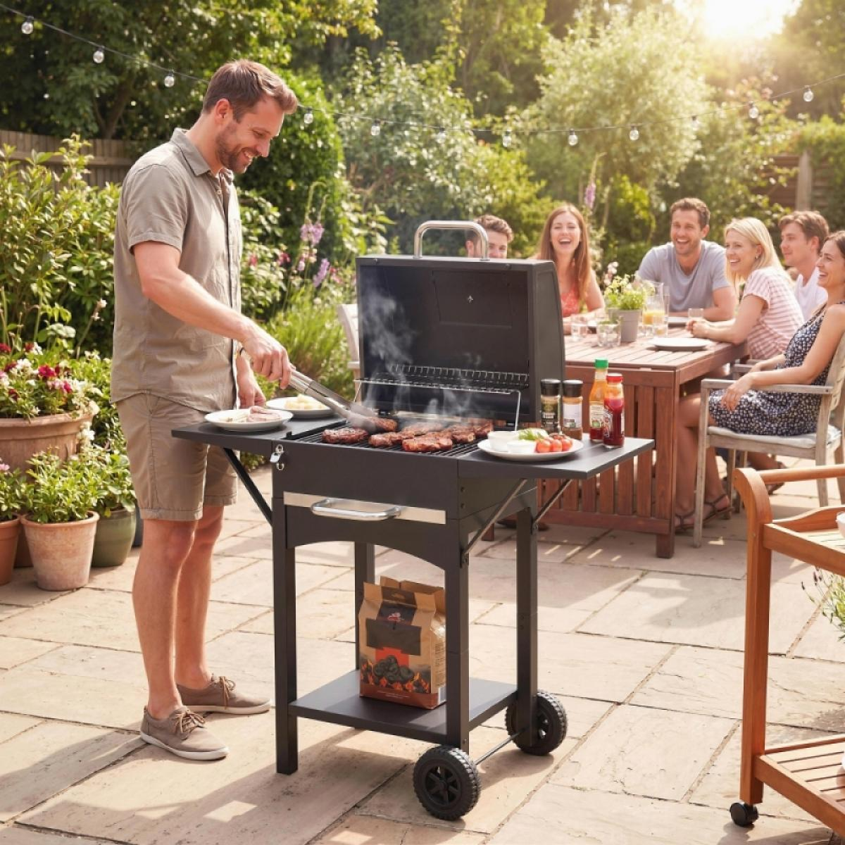 Mann grillt Fleisch auf einem Grill, während Freunde am Tisch sitzen.