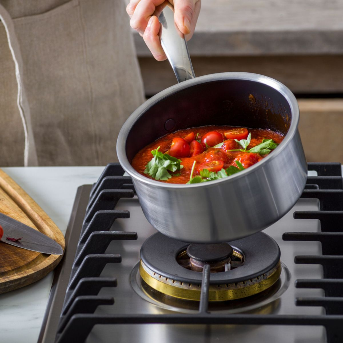 Una persona sostiene una cacerola plateada llena de salsa de tomate roja, tomates cherry y hierbas verdes sobre una estufa de gas.