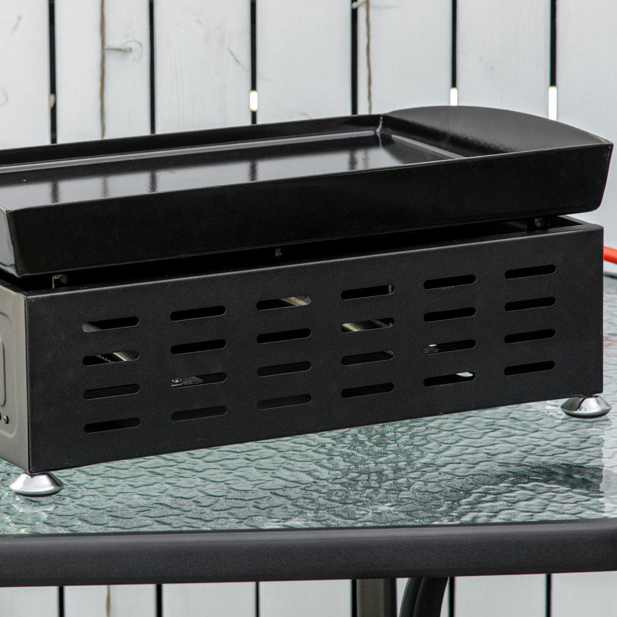 Eine schwarze Grillplatte steht auf einem Glastisch. Die Platte hat eine flache Oberseite und einen belüfteten Sockel.