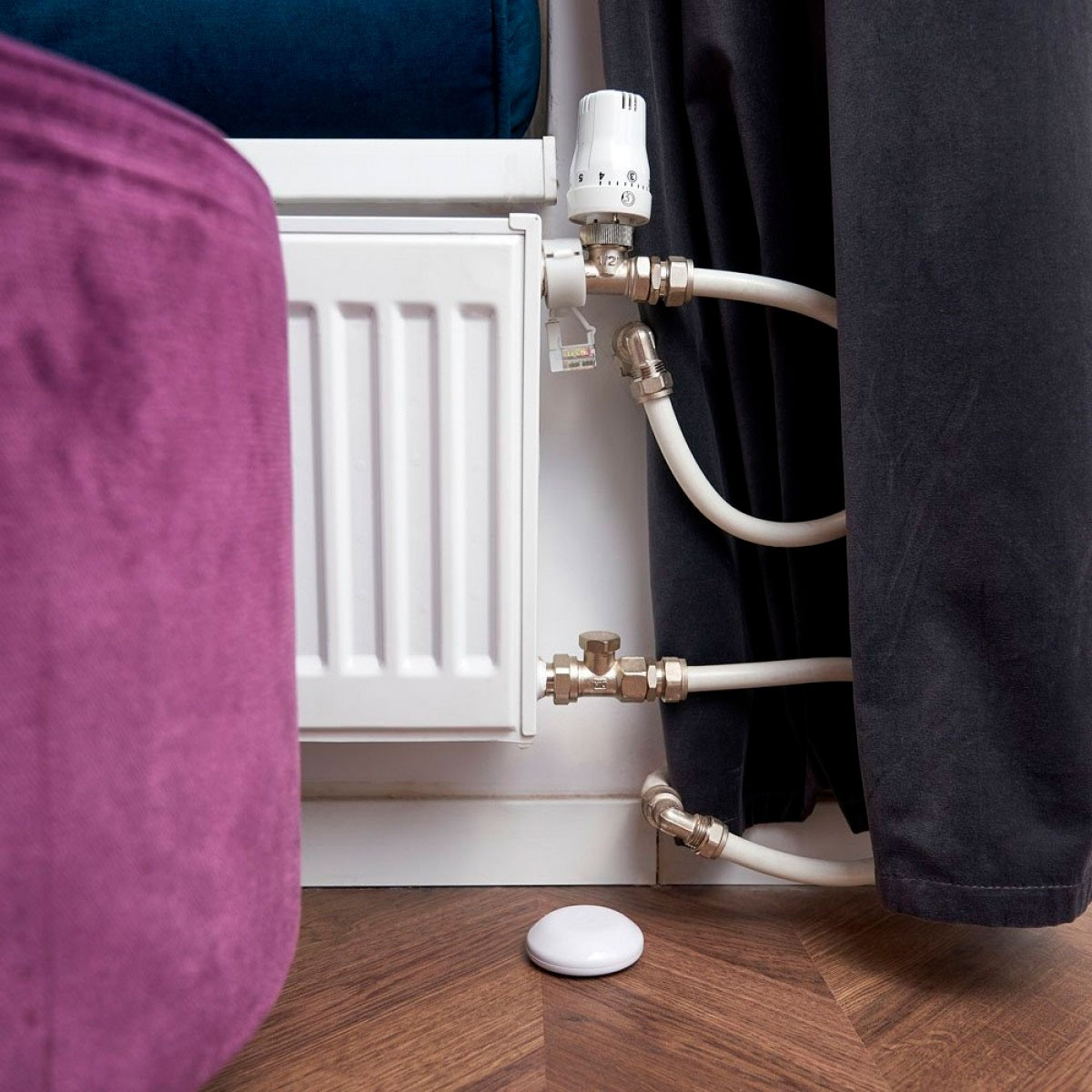 Radiatore bianco con valvola termostatica e tubi, accanto a un pouf viola e una tenda scura.