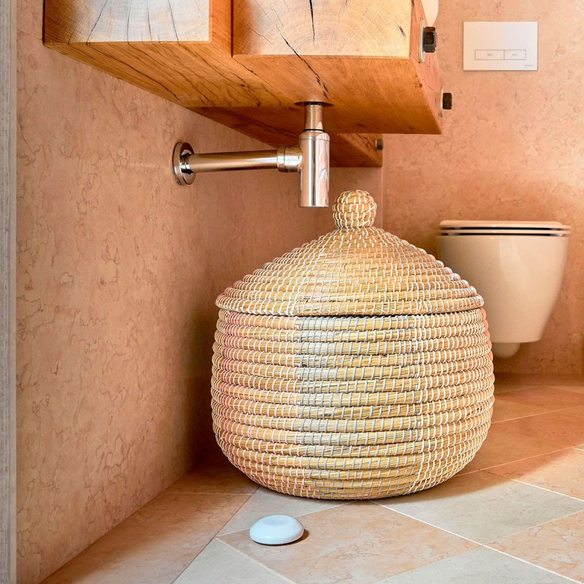 Un cesto rotondo in vimini con coperchio sotto un lavandino, vicino a un WC.
