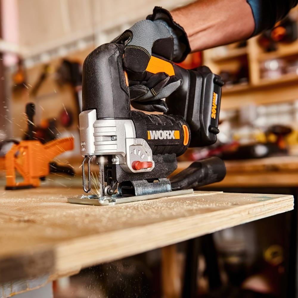Eine behandschuhte Hand bedient eine schwarz-orange Stichsäge, die ein Holzbrett schneidet. Sägemehl fliegt.