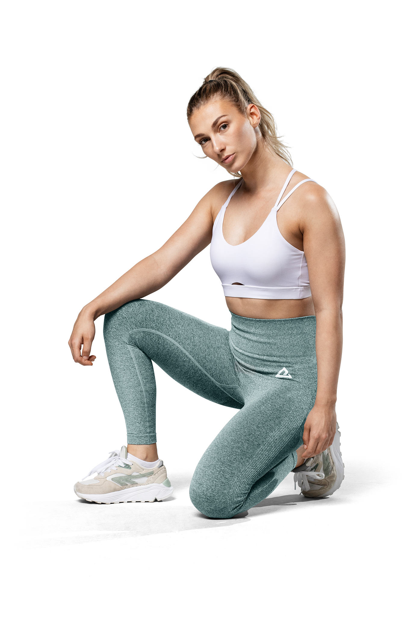 Una mujer con ropa deportiva se arrodilla, top blanco, leggings verdes, zapatillas blancas.
