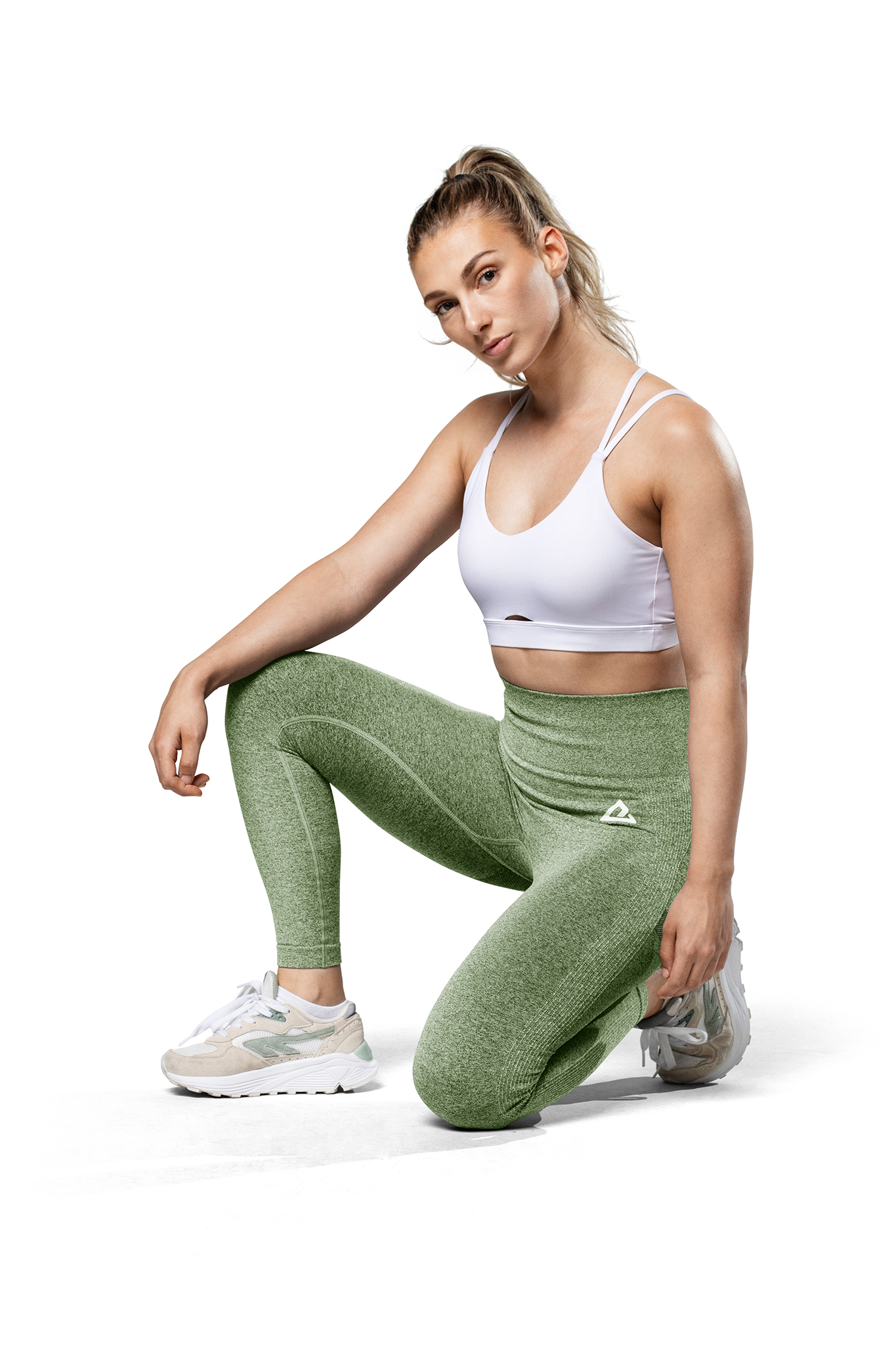 Mujer con ropa deportiva arrodillada sobre fondo blanco, leggings verdes y sujetador blanco.