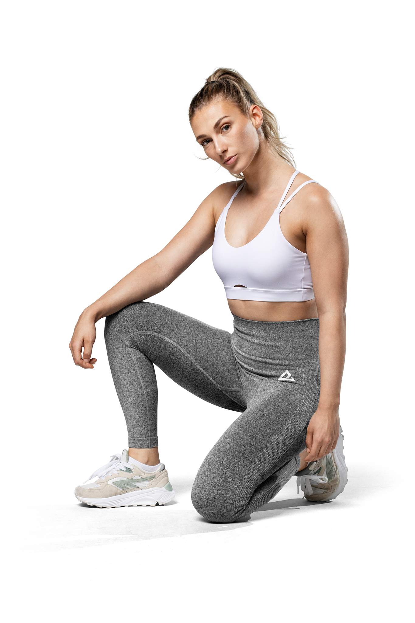 Una mujer con sujetador deportivo blanco y leggings grises se arrodilla.