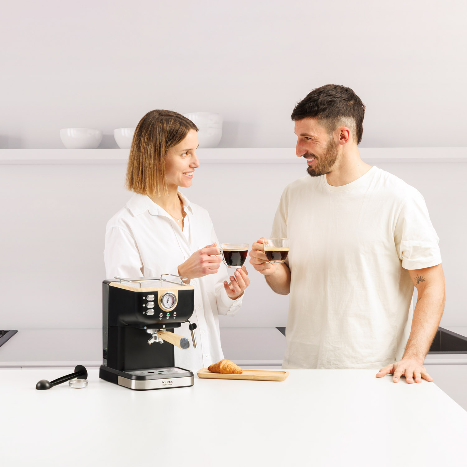 Hombre y mujer brindan con café en cocina luminosa. Cafetera y croissant.