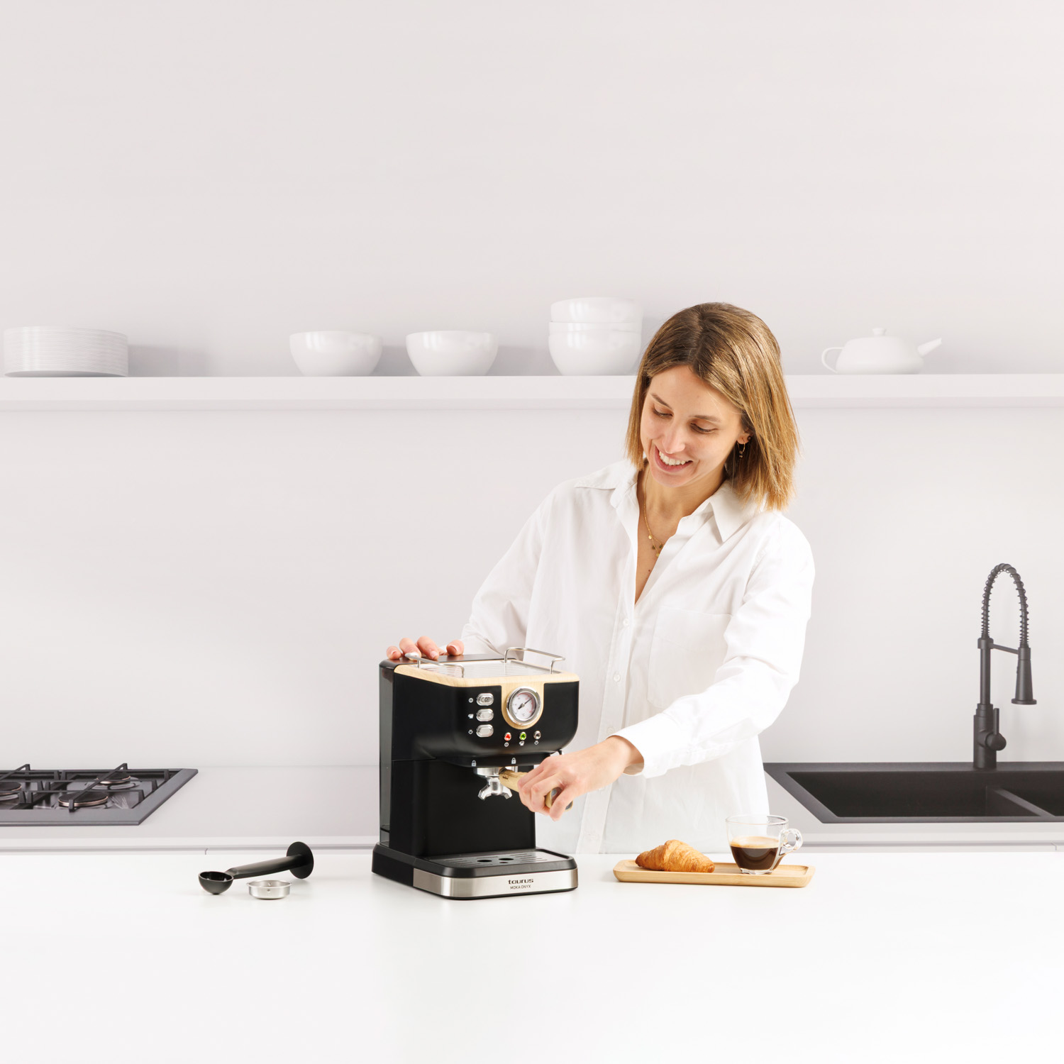 Mujer con camisa blanca prepara café con máquina, croissant y café cerca.