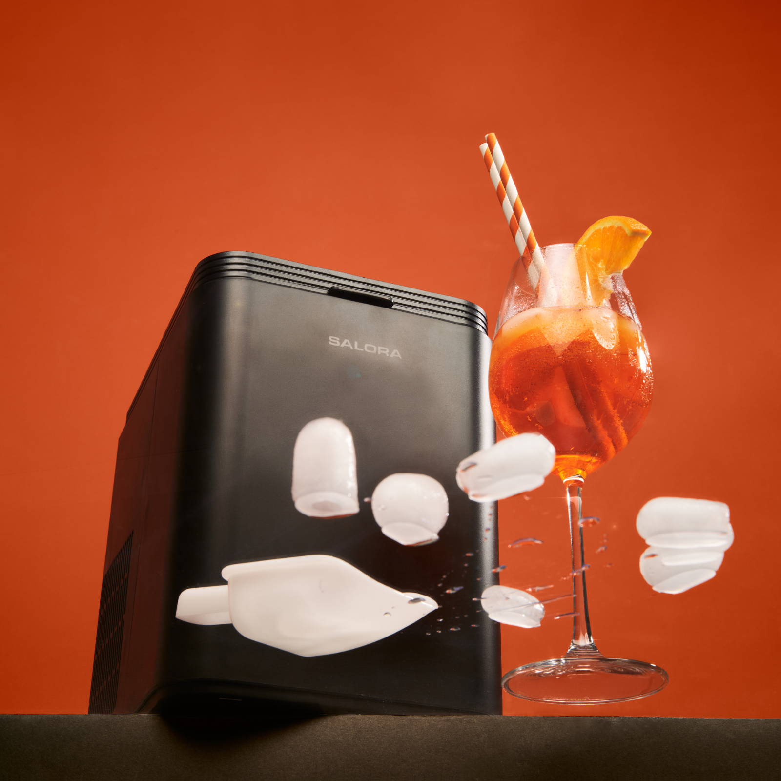 Una máquina de hielo negra con un vaso de Aperol Spritz y cubitos de hielo volando.