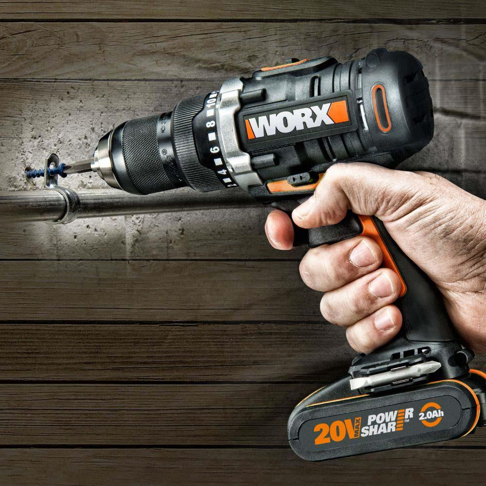 Una mano tiene un trapano Worx nero e arancione, che fora una vite blu in un muro di legno.
