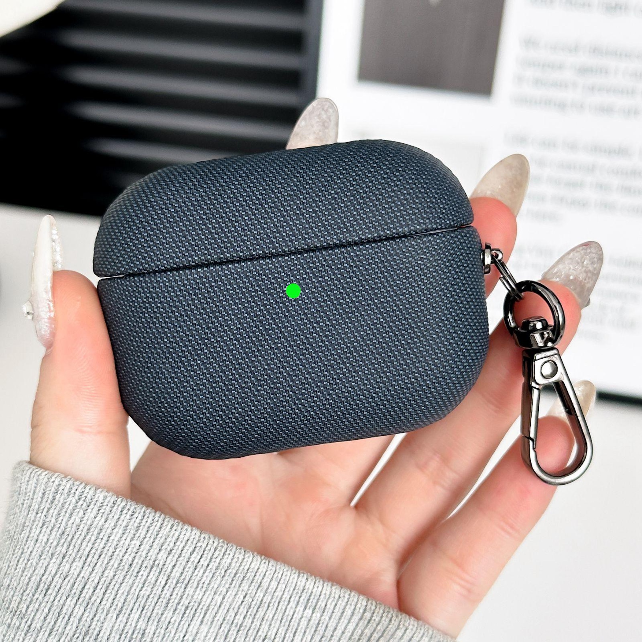 Funda gris oscura para AirPod con clip de mosquetón, sostenida por una mano con esmalte.