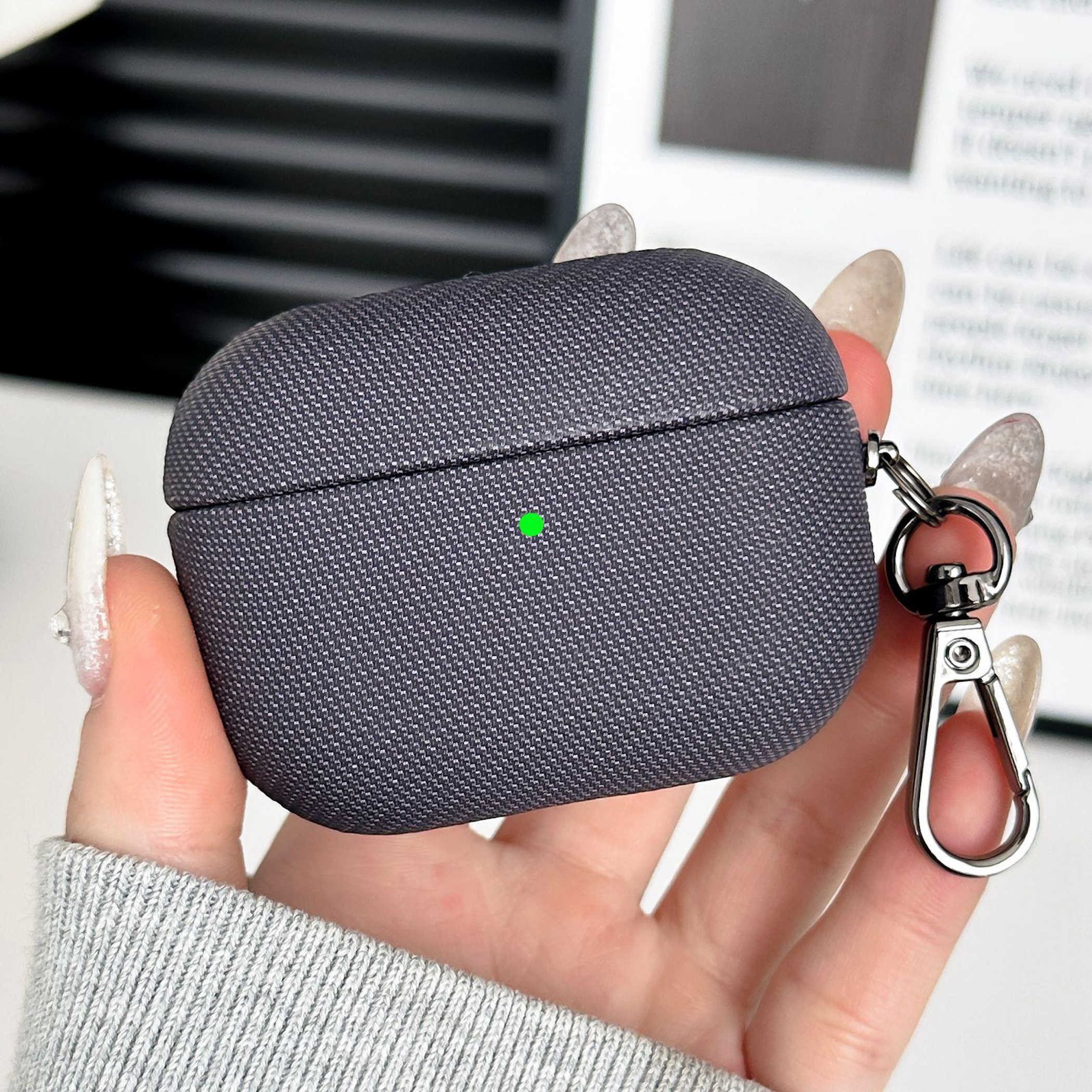 Eine Hand hält eine graue AirPods Pro Hülle mit einem Schlüsselring. Ein grüner Punkt ist sichtbar.