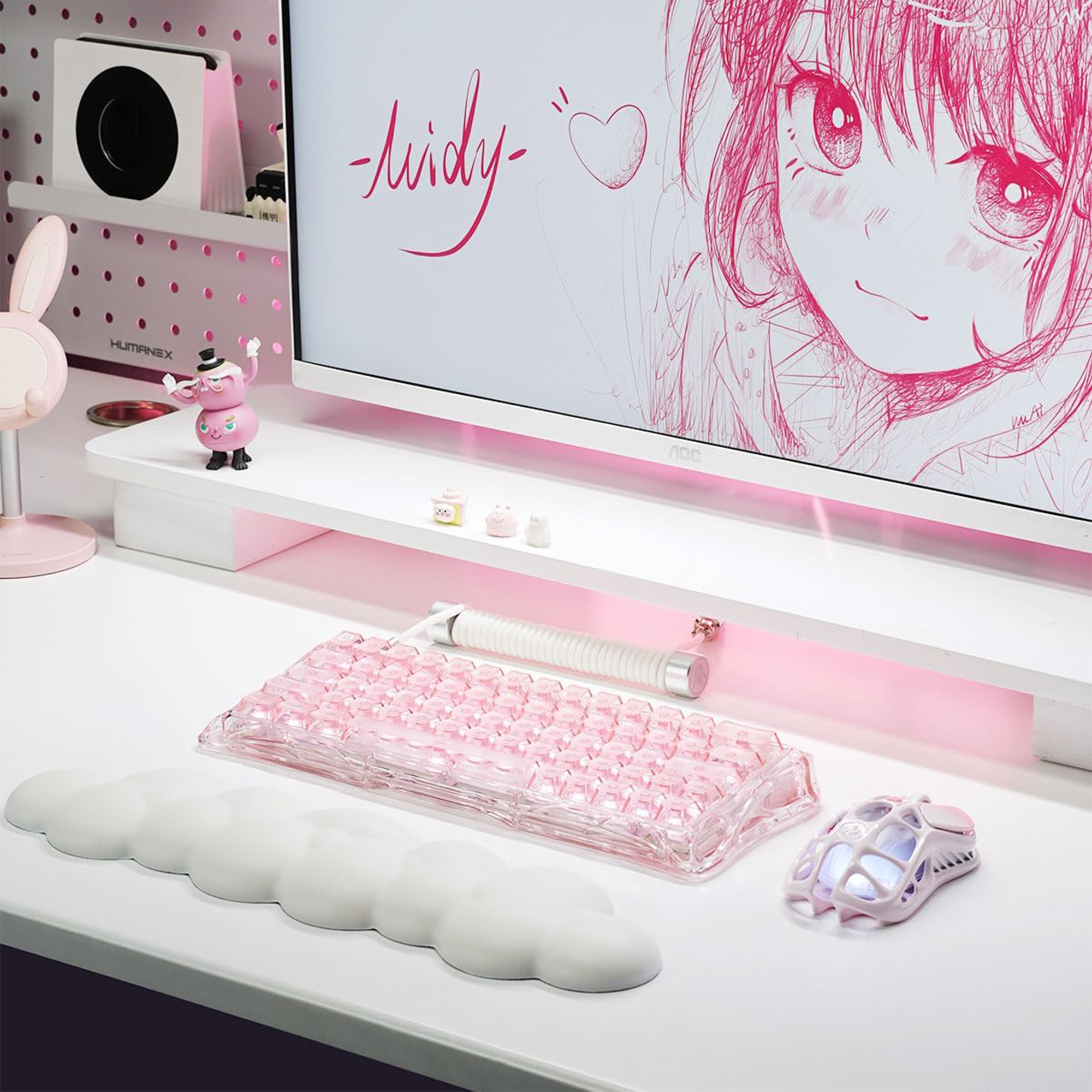Monitörde klavye, fare, figür ve anime çizimi bulunan pembe bir oyuncu masası.