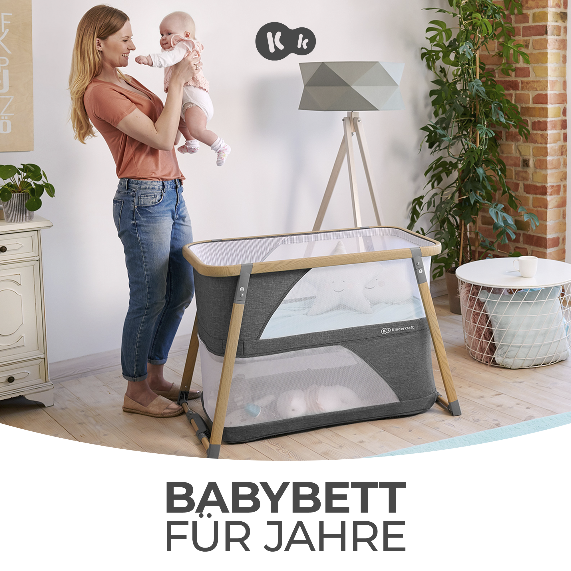 Eine Frau hält ein Baby vor einem Kinderbett mit Holzrahmen und grauen Stoffseiten.
