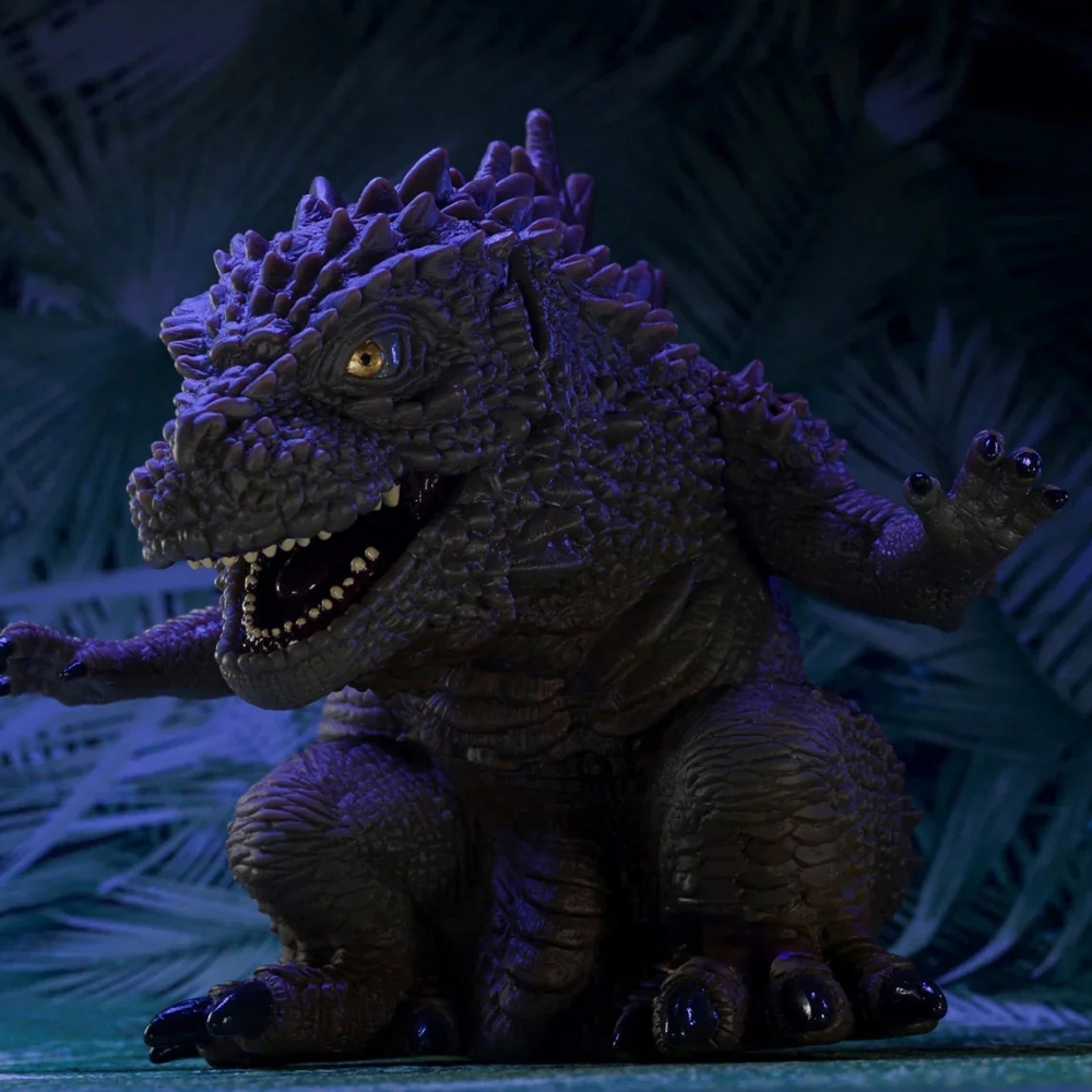 Eine dunkle Godzilla-Figur sitzt mit offenem Mund und gelben Augen vor einem tropischen Hintergrund.