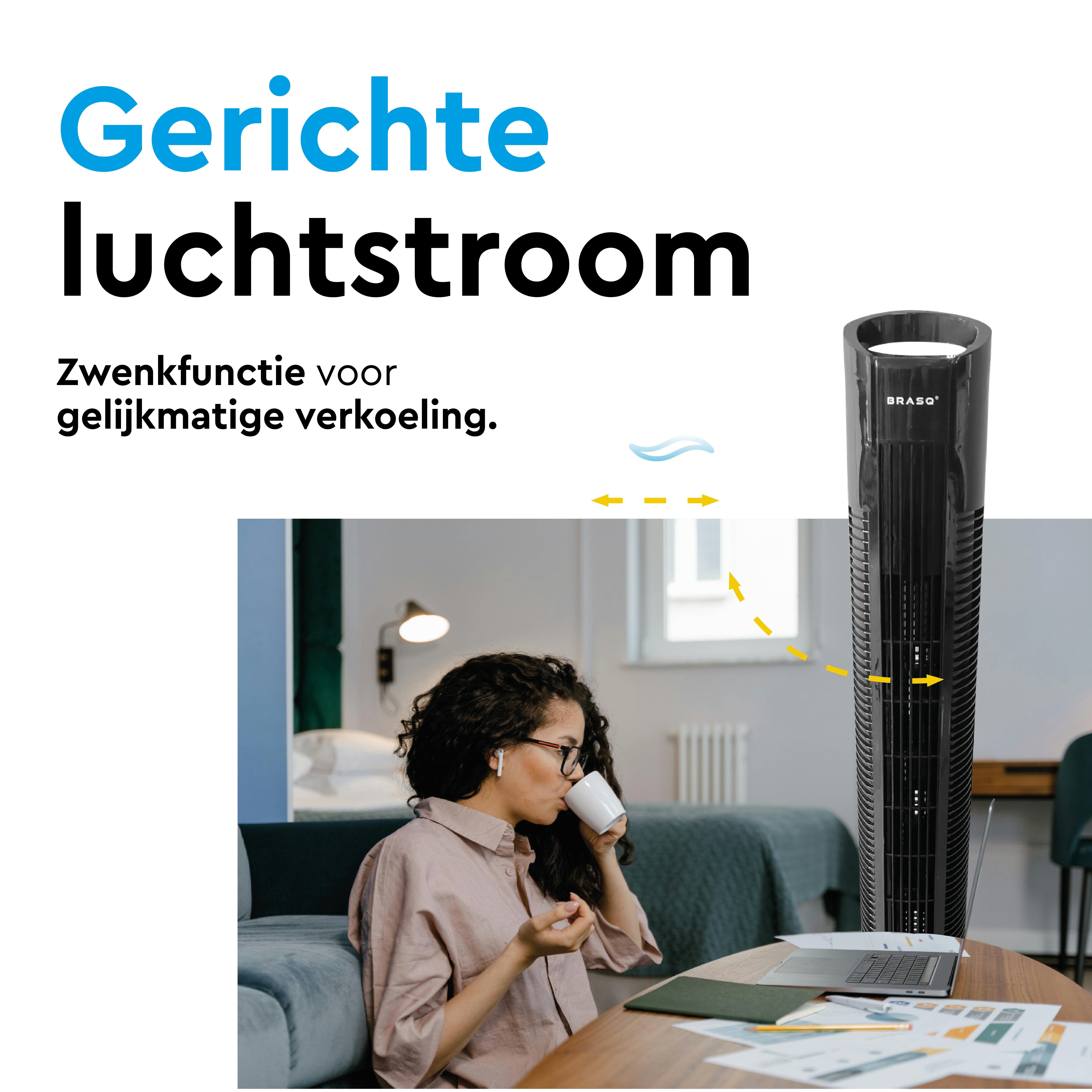 A slim black tower fan is next to a laptop and papers. A woman drinks coffee.