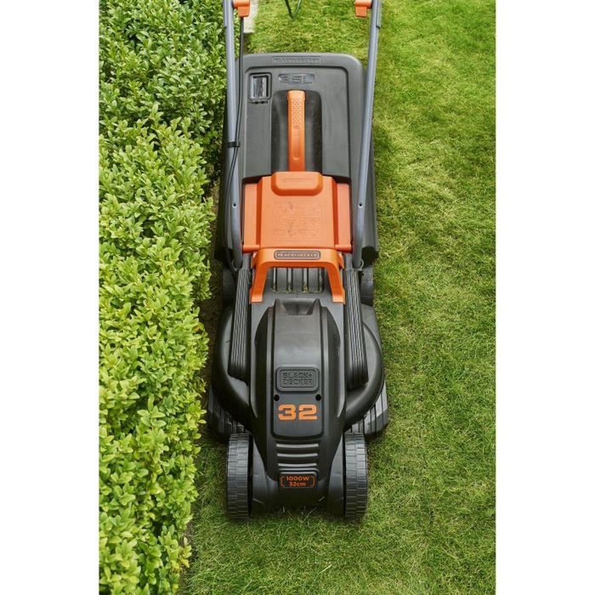 Un cortacésped negro con detalles naranjas se asienta sobre un césped verde junto a un seto.