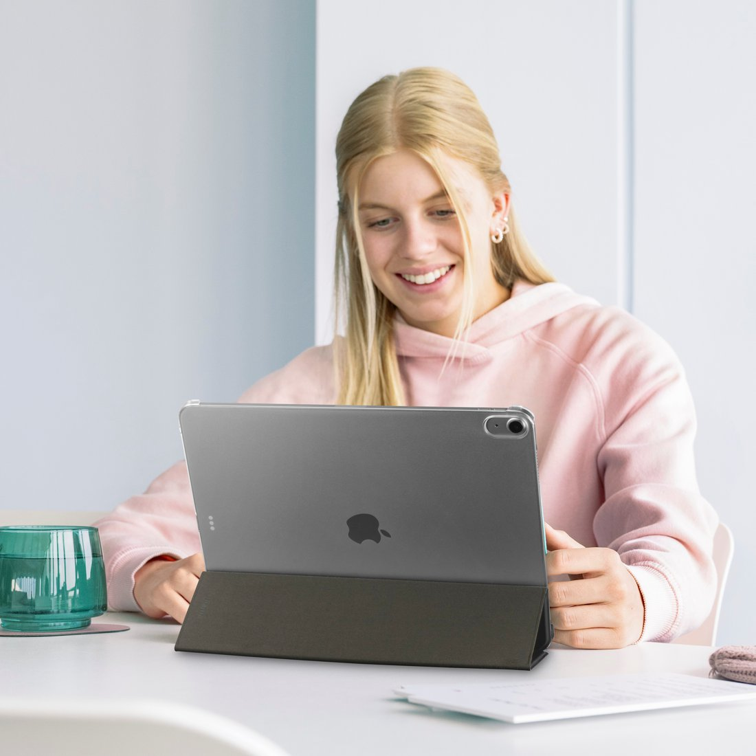 Blondynka w różowej bluzie z kapturem uśmiecha się, używając tabletu na białym stole.