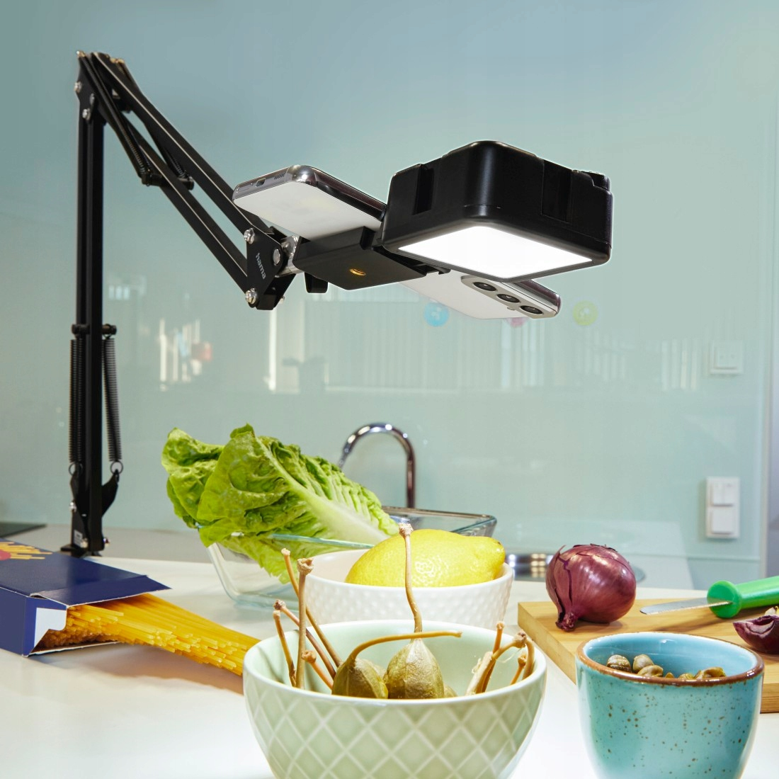 Smartfon i lampa na regulowanym ramieniu nad blatem kuchennym ze spaghetti, sałatą, cytryną, kaparami, cebulą i orzechami.