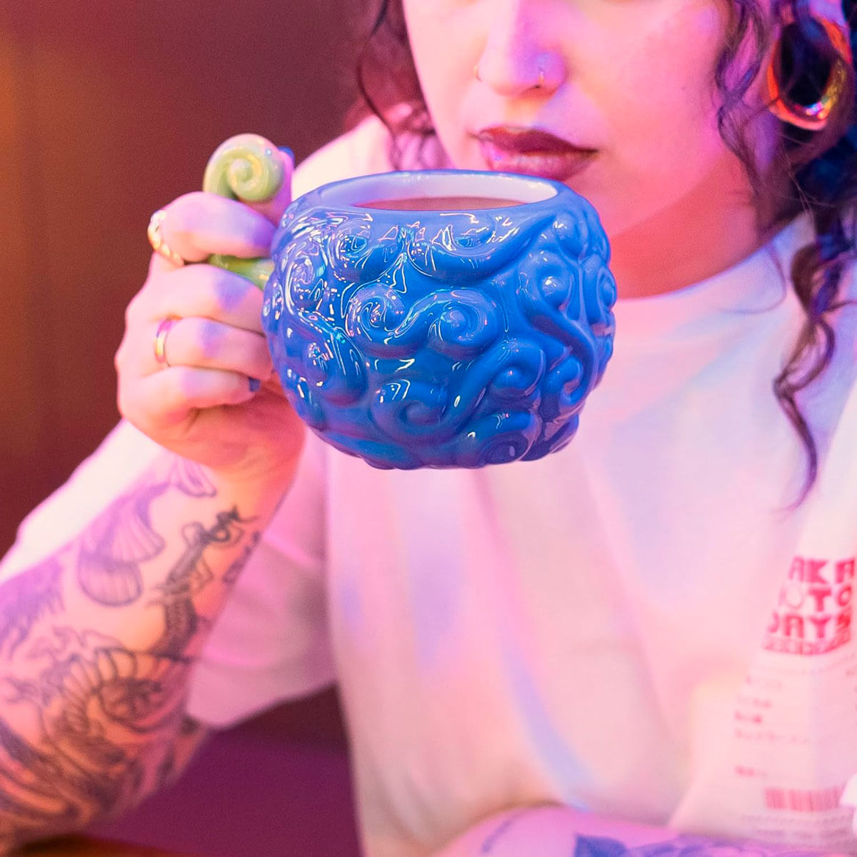 Una persona tatuada sostiene una taza azul con forma de cerebro y asa verde.