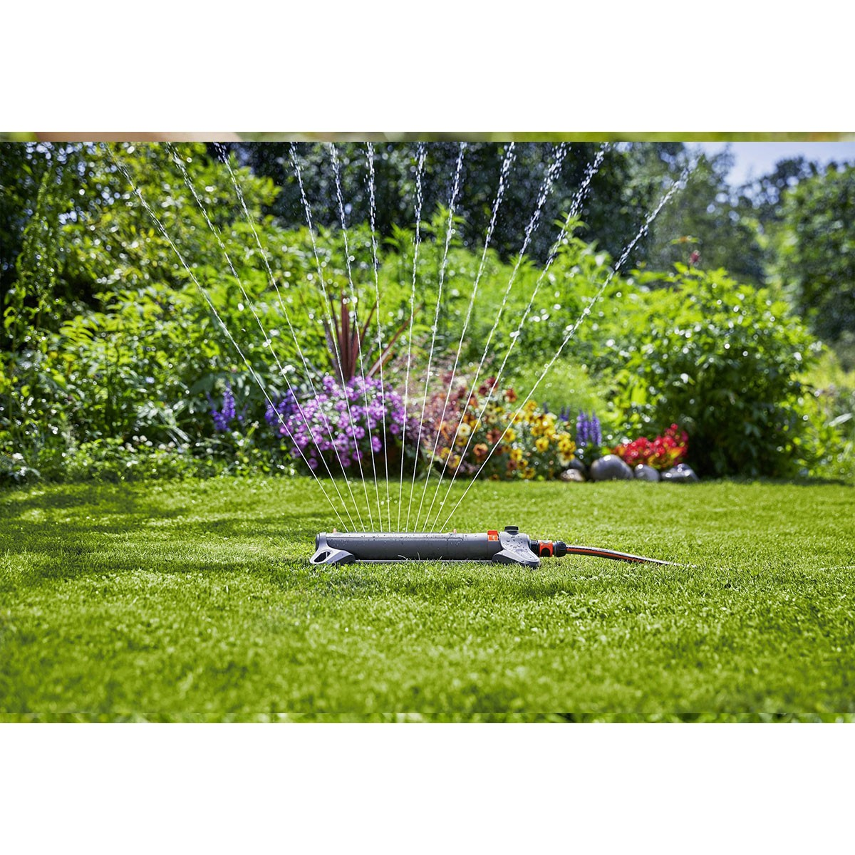 Un irrigatore da giardino spruzza acqua su un prato verde lussureggiante con fiori colorati sullo sfondo.