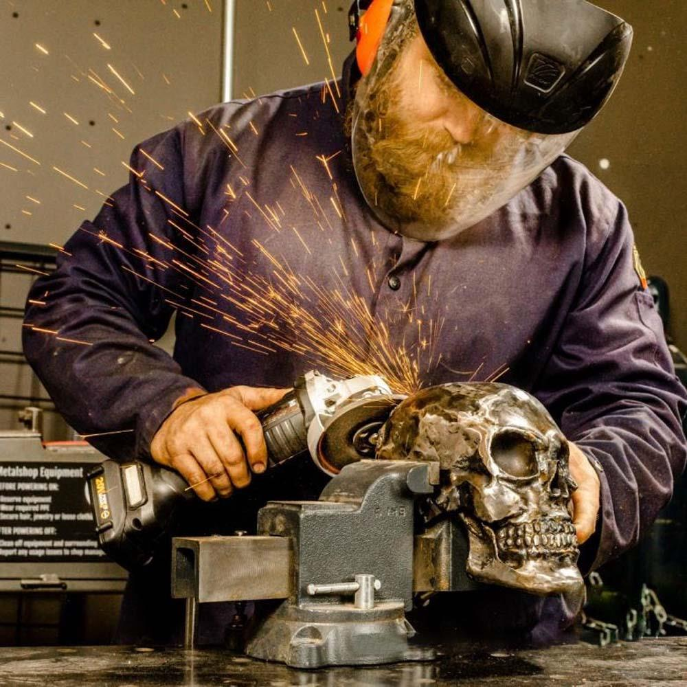 Güvenlik teçhizatlı adam mengenedeki metal kafatasını taşlıyor, kıvılcımlar uçuşuyor.