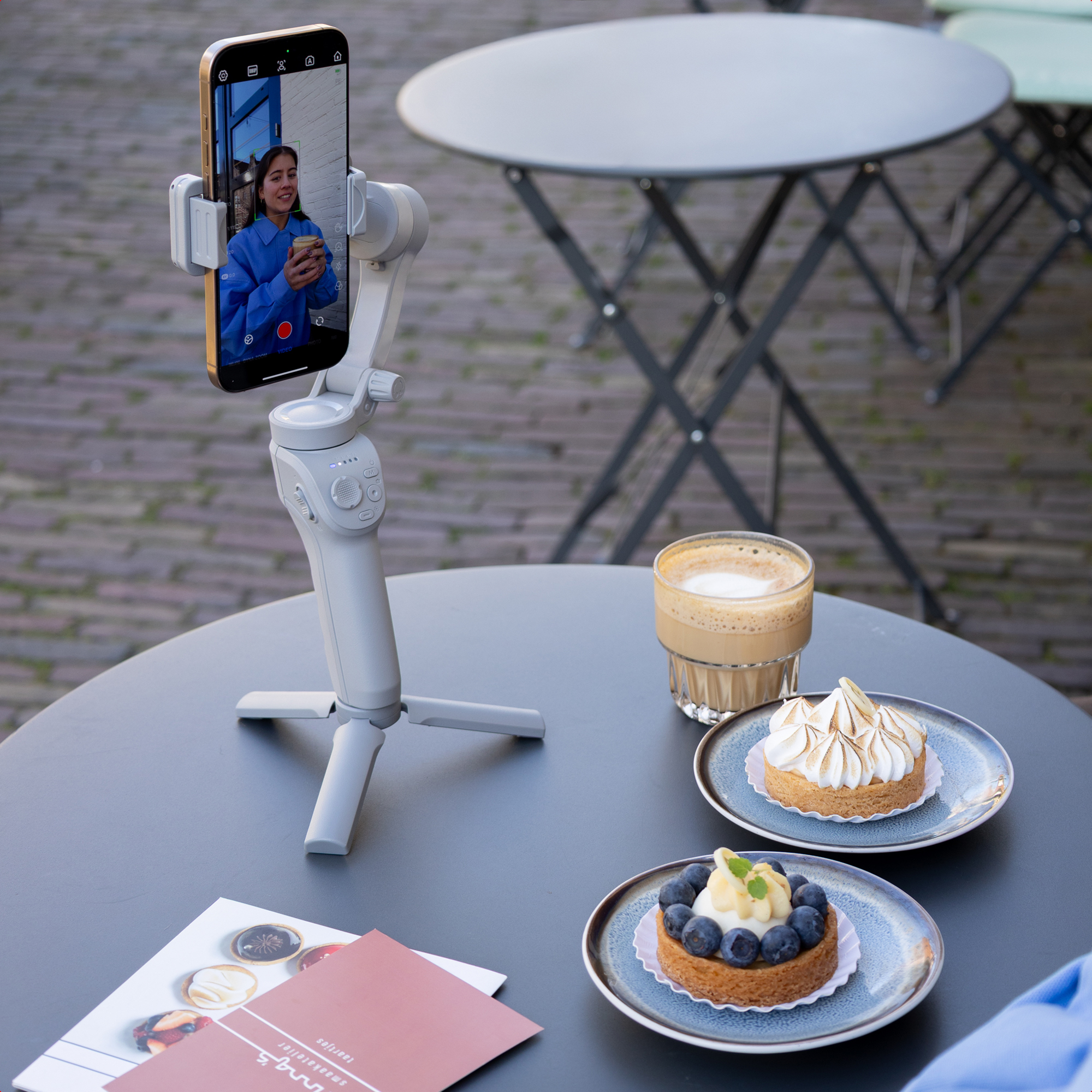 A gimbal holds a phone showing a woman with coffee. Two tarts and coffee on a table.