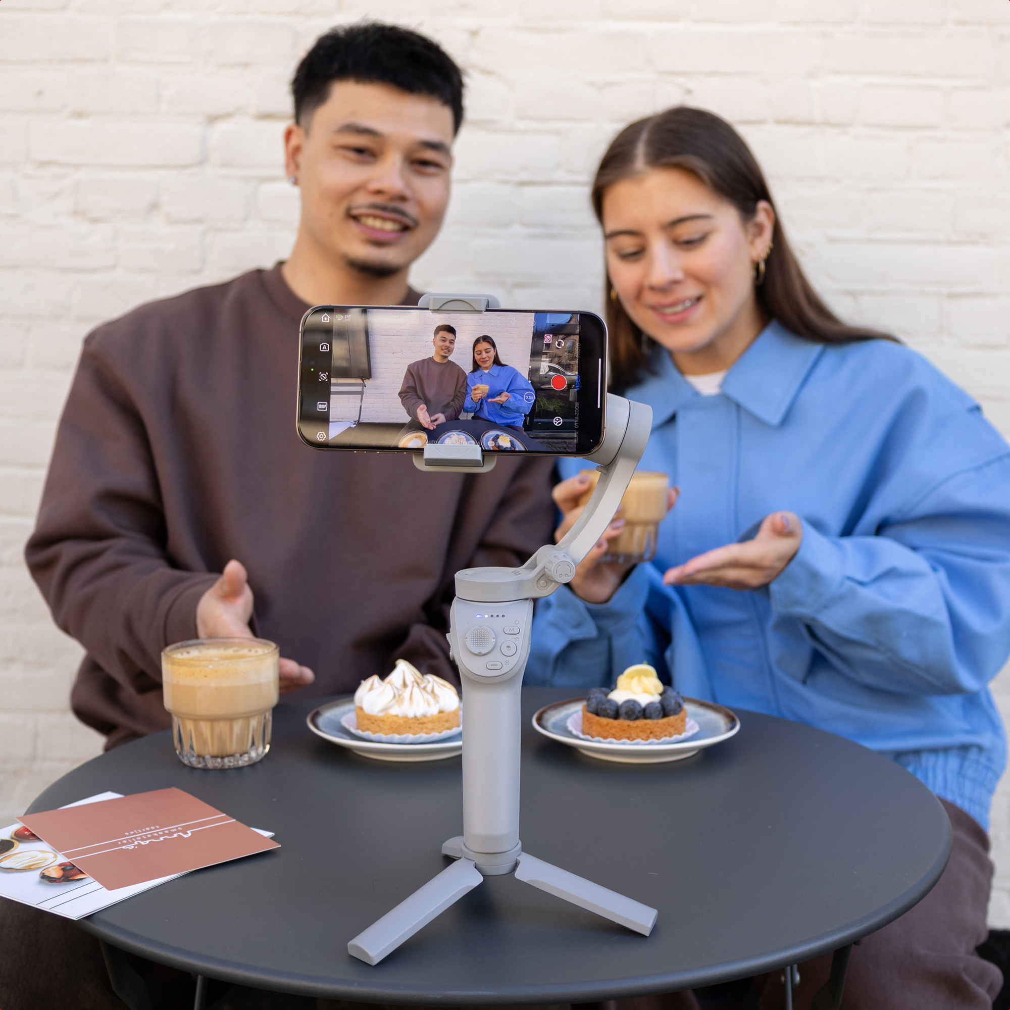 A man and woman sit at a table with coffee and cakes, filming themselves with a gimbal.