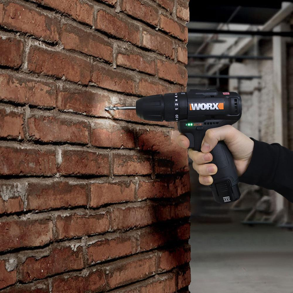 Una mano tiene un trapano Worx nero, che fora una parete di mattoni rossi. Il trapano ha una punta argentata.