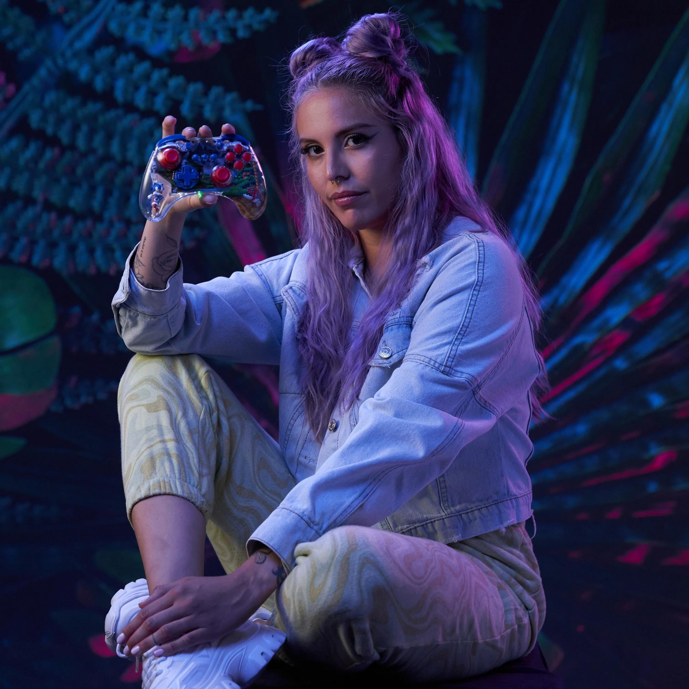 Frau mit lila Haaren hält einen Controller mit Sonic-Motiv vor einem dunklen Hintergrund mit Pflanzen.