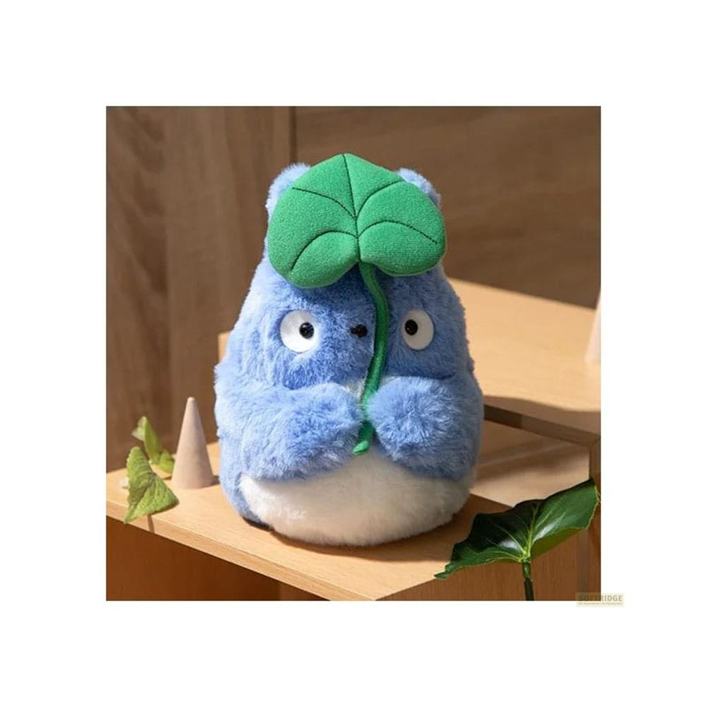 Un Totoro en peluche bleu tenant une feuille verte. Arbres et bois en arrière-plan.
