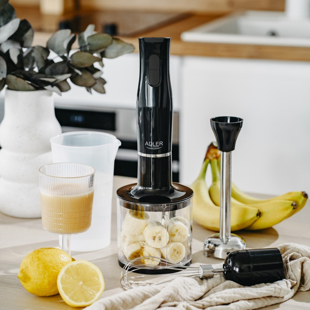 Czarny blender ręczny z akcesoriami, bananami, cytryną i szklanką soku na drewnianym stole.