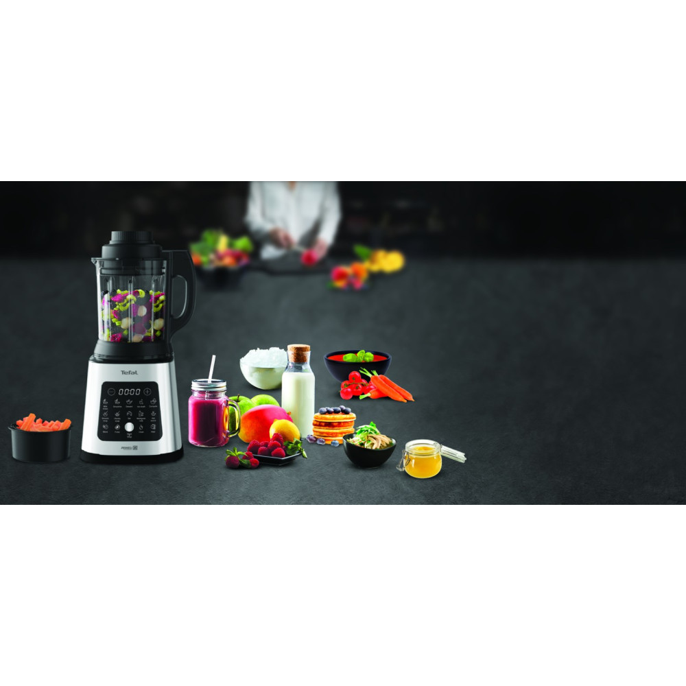 Ein Mixer mit Obst, Gemüse und Beilagen auf einem dunklen Tisch.