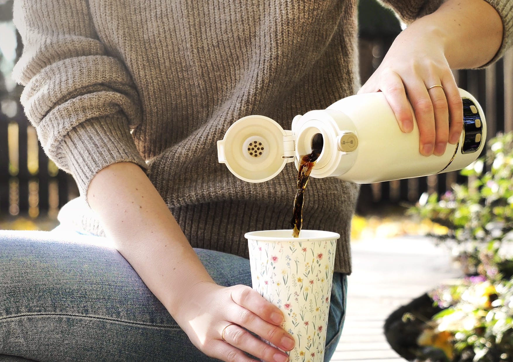Eine Person gießt Kaffee aus einer Thermoskanne in eine gemusterte Tasse.