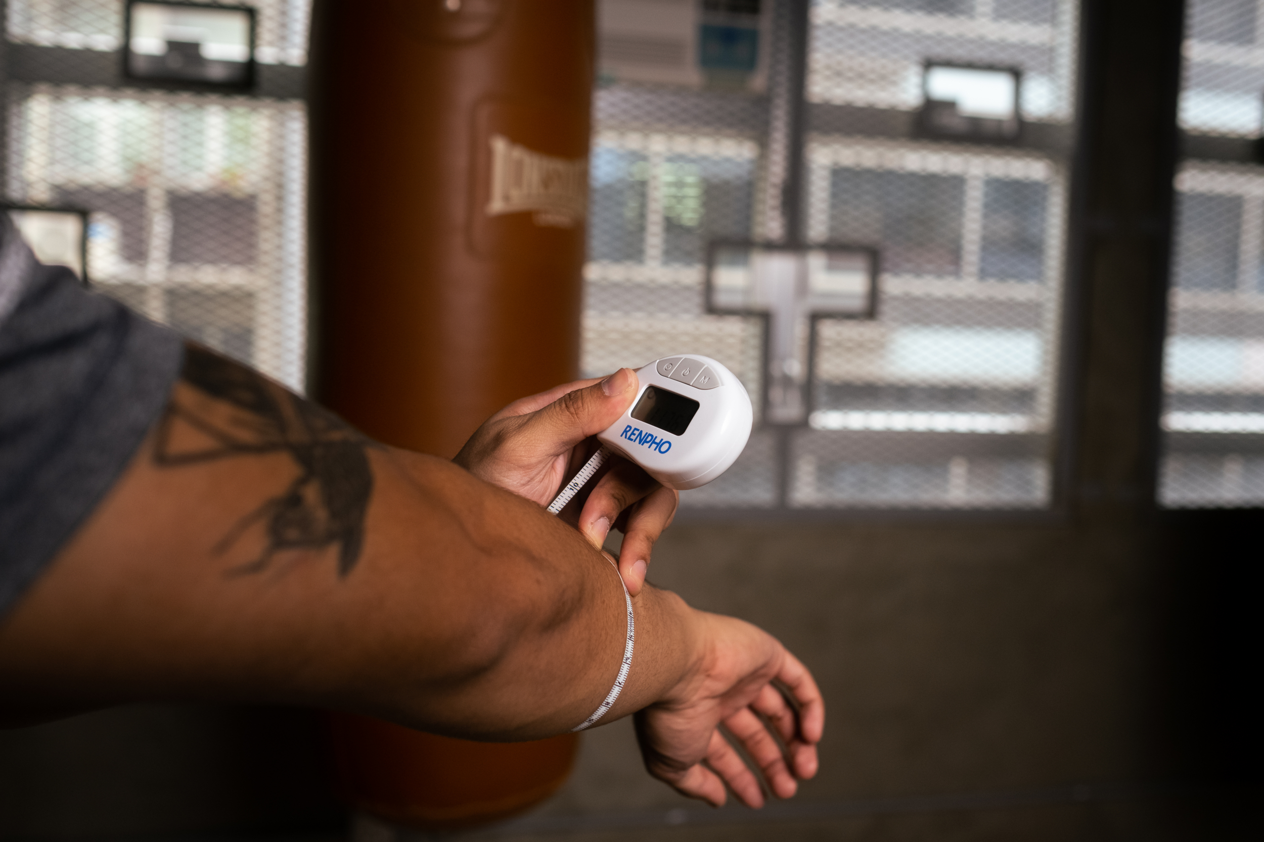 Una mano mide un brazo con una cinta métrica blanca. Un saco de boxeo está al fondo.