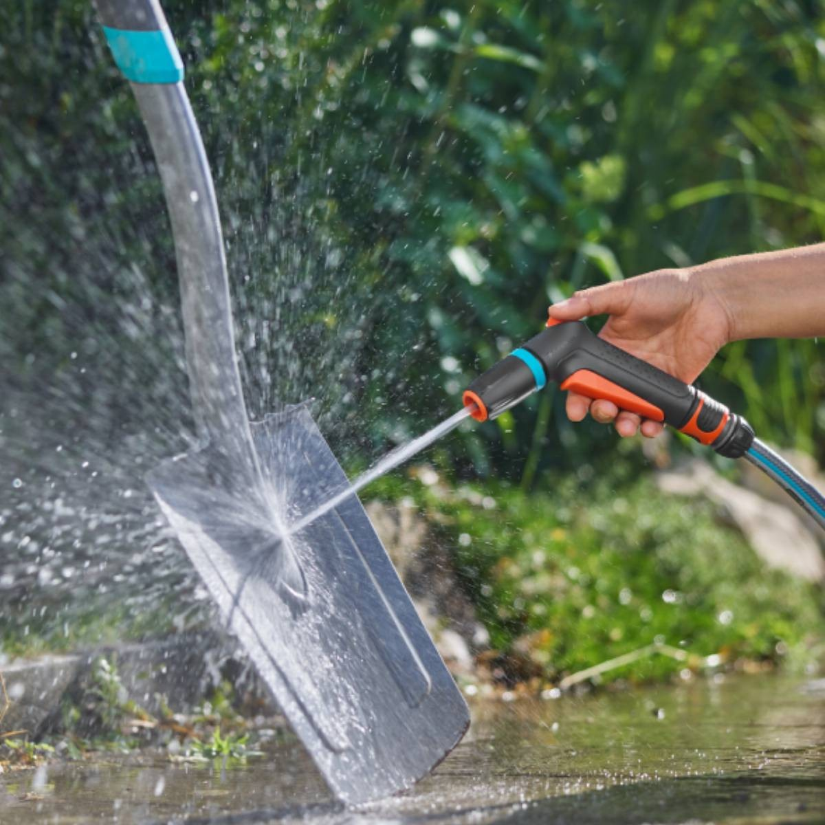Una mano spruzza acqua da un tubo su una pala. Fogliame verde sullo sfondo.