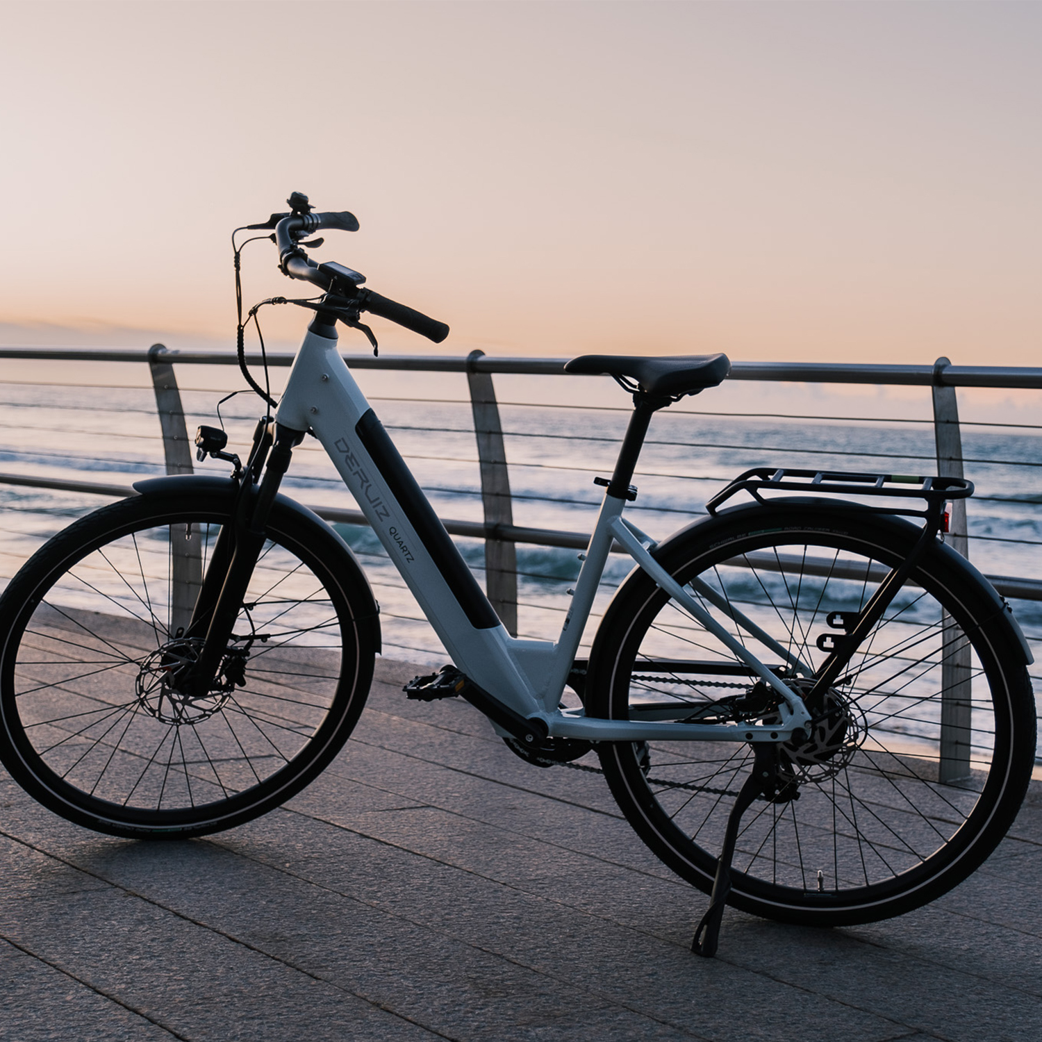 Ein weißes Elektrofahrrad steht auf einem Gehweg am Meer bei Sonnenuntergang.