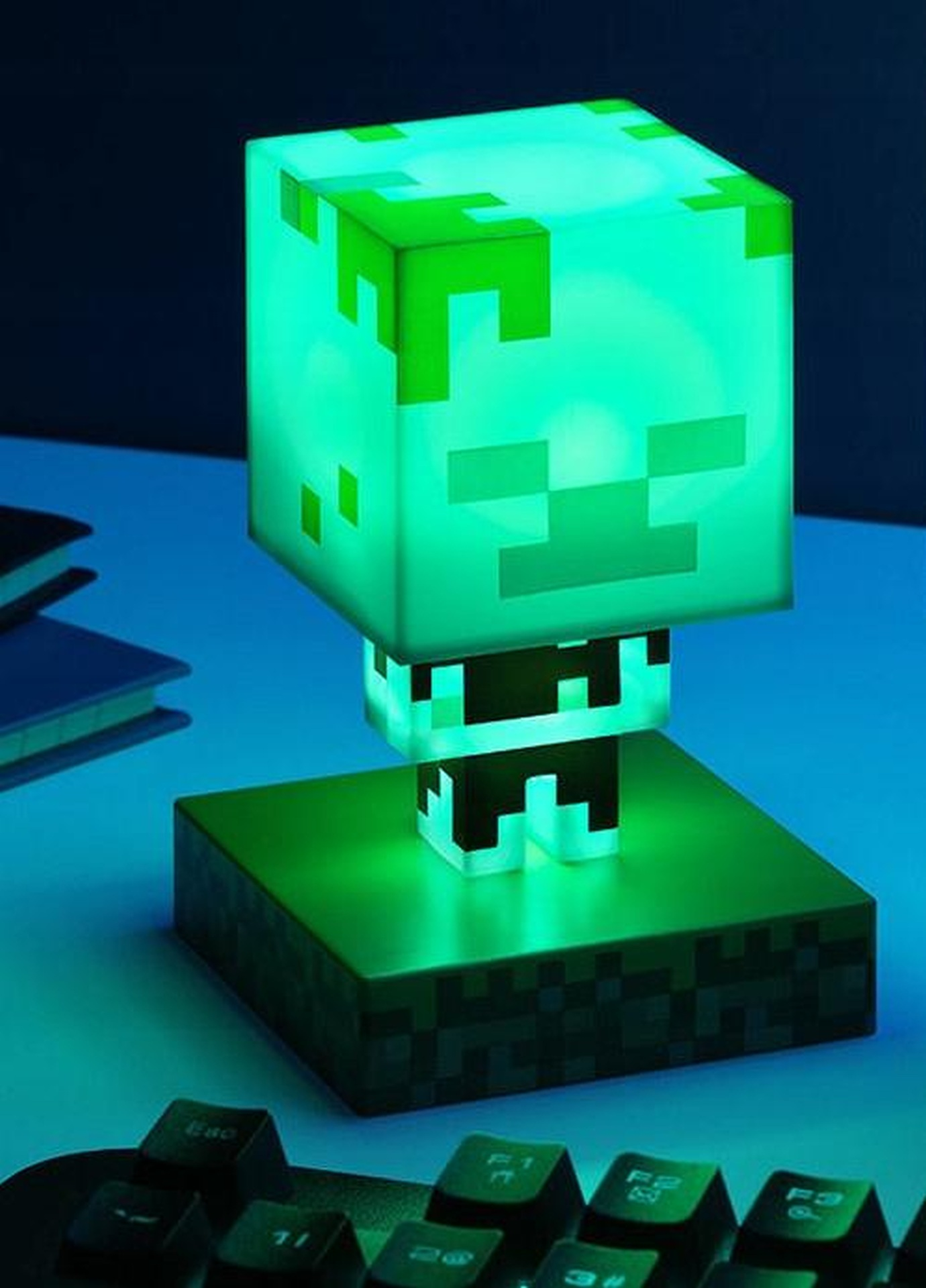 Zielona lampka Minecraft Creeper świeci na biurku obok książek i klawiatury.