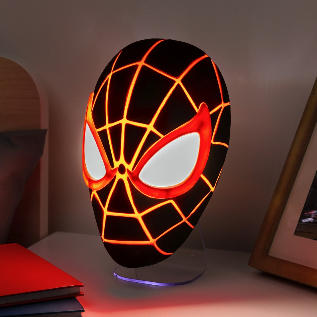 Lampa w kształcie maski Spidermana świeci na szafce nocnej z książkami i ramką.