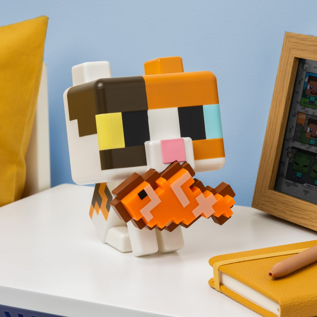 Pikselowa zabawka kota Minecraft trzyma pomarańczową rybę. Widoczne są żółta poduszka i oprawiona grafika.