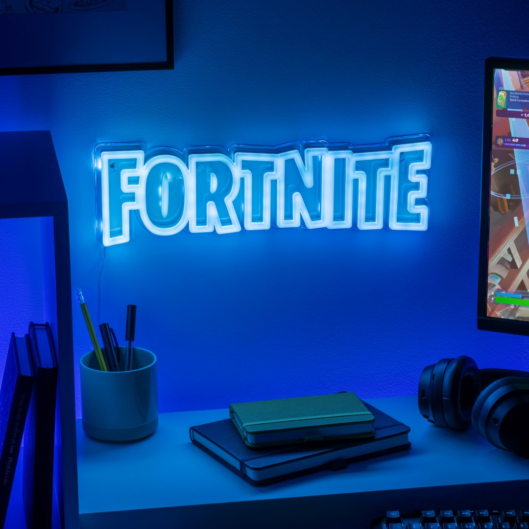 Niebieski neonowy znak Fortnite świeci nad biurkiem z monitorem, słuchawkami i notatnikami.