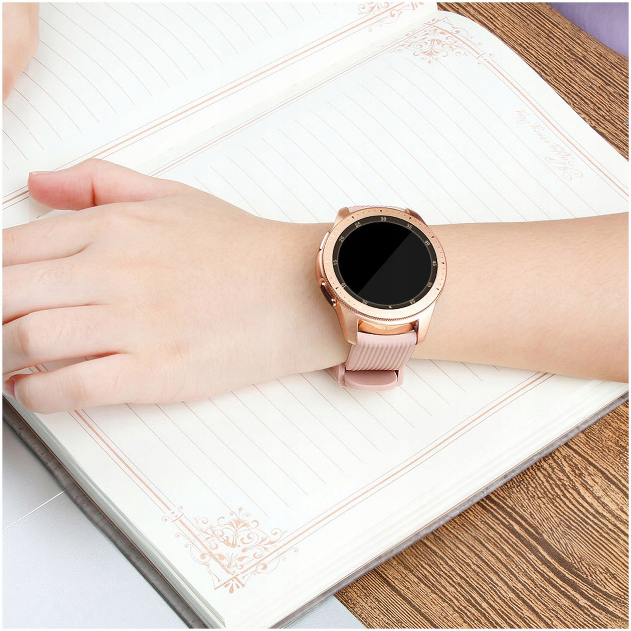 Różowo-złoty smartwatch z różową bransoletą na nadgarstku osoby, na otwartym notatniku.