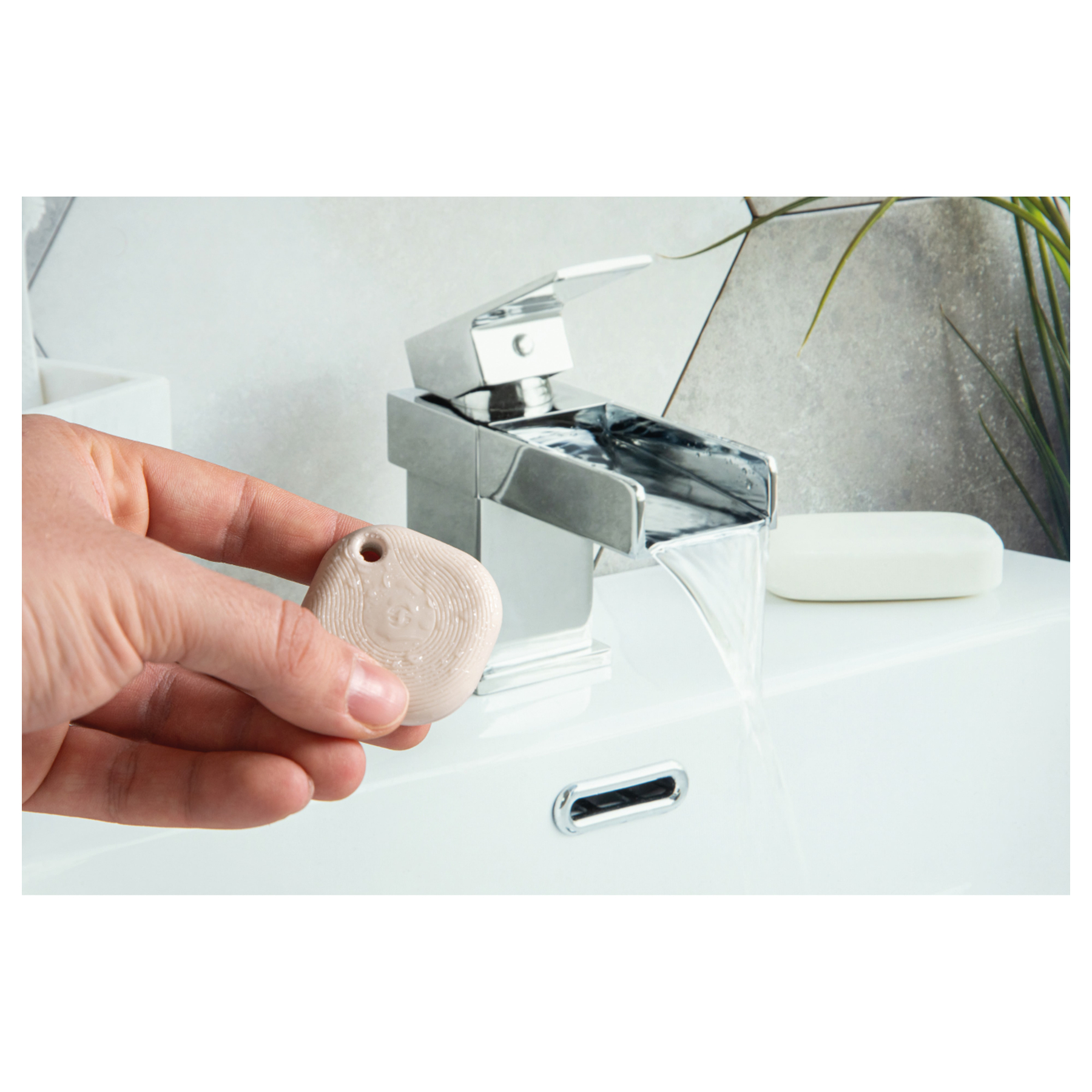 Une main tient un pendentif beige clair au-dessus d'un lavabo avec de l'eau courante.